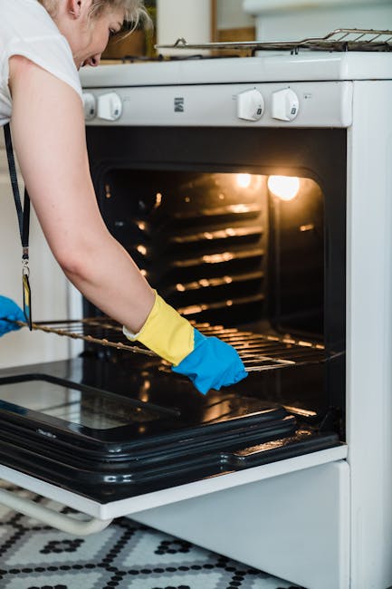 A close-up view inside an oven showcasing a black ceramic or enamel surface with soap suds and water droplets. A person wearing blue rubber gloves and a yellow sleeve is wiping the surface with a pink